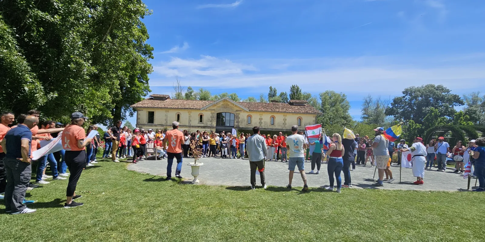 Large outdoor community gathering in front of a historic beige stone building with many participants wearing orange shirts, holding various flags, on a sunny day in Bordeaux.