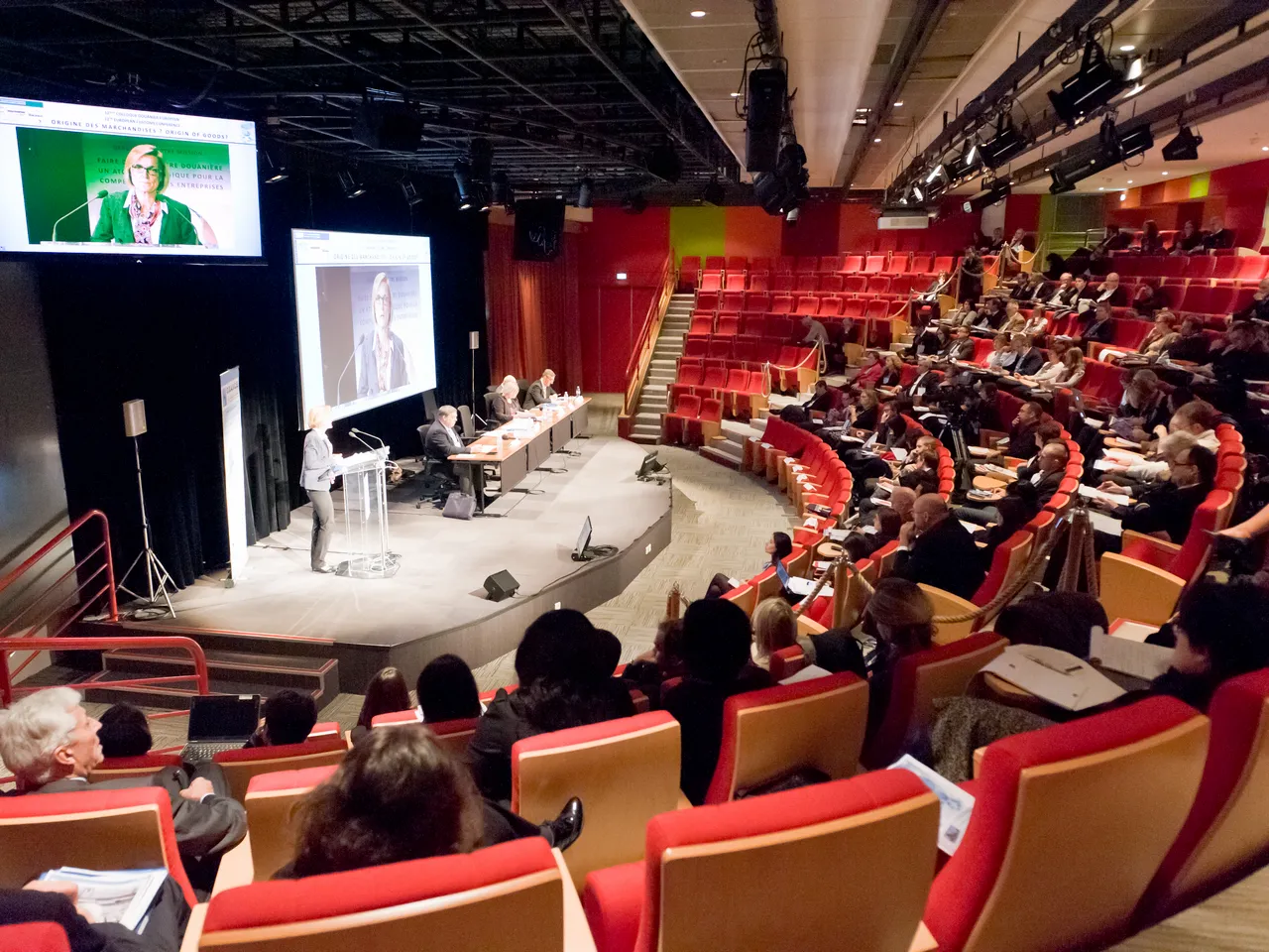 A conference hall at Cité Mondiale in Bordeaux with an audience seated in red and tan chairs watching a panel of speakers on stage, with projection screens displaying presentations.