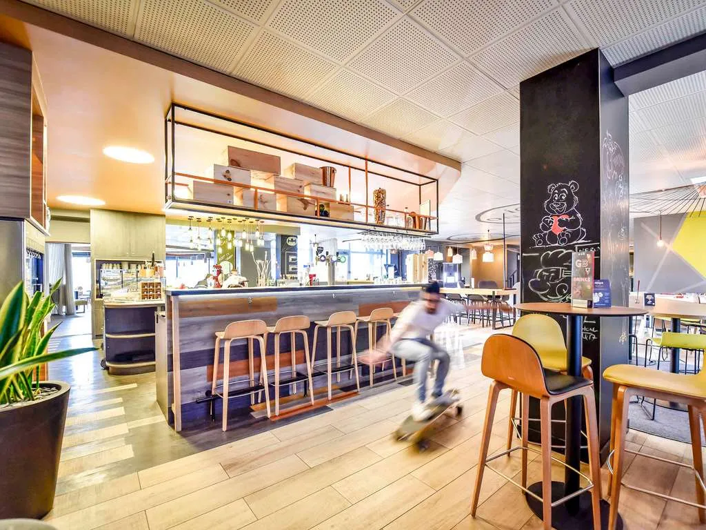 Modern hotel bar and lounge area at Novotel Bordeaux Lac with wooden bar stools, contemporary furniture, open kitchen, and a blurred figure in motion