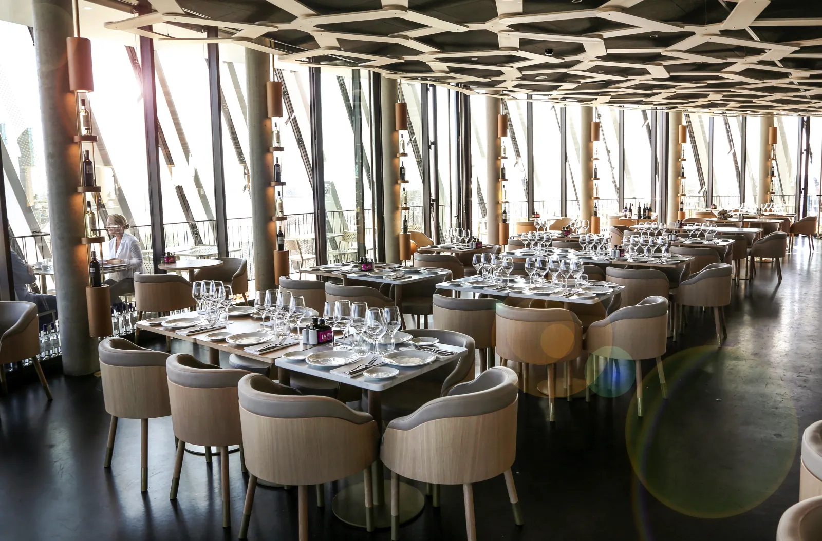Modern panoramic restaurant interior with contemporary seating, tables set for dining, floor-to-ceiling windows overlooking Bordeaux cityscape, geometric ceiling design, and neutral-toned furnishings.
