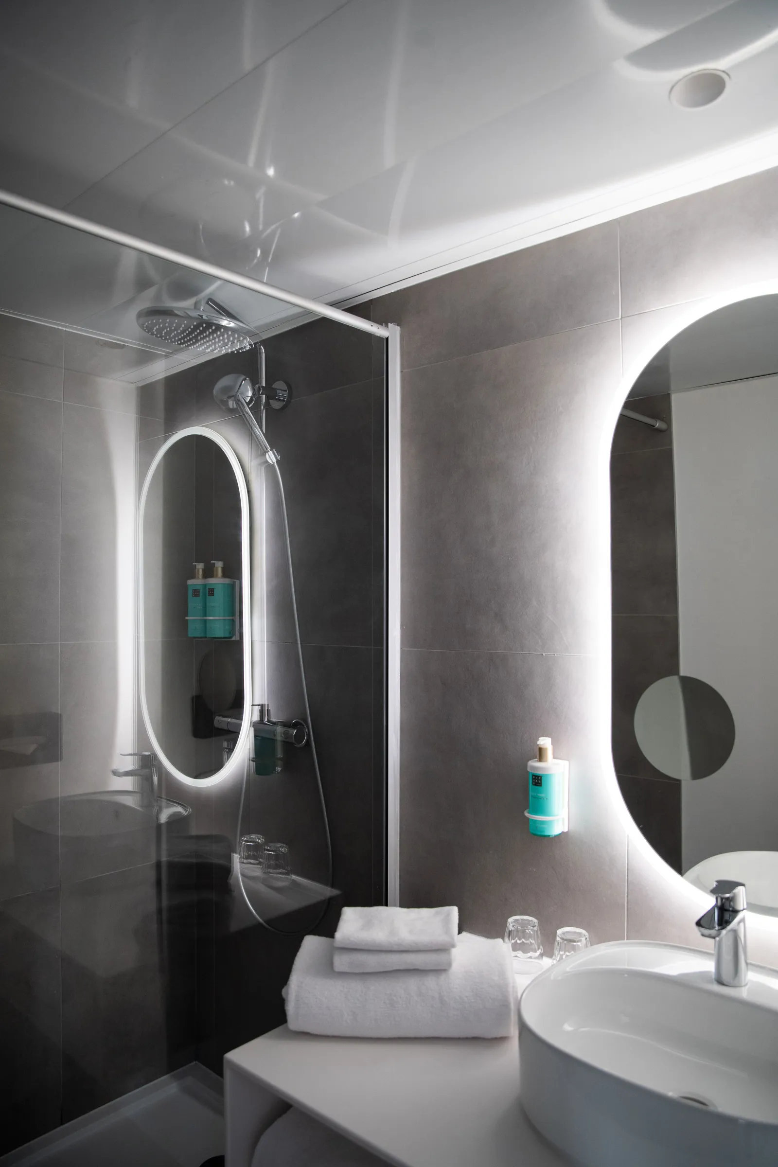 Modern bathroom with illuminated oval mirror, black and white tile walls, round white sink, rainfall showerhead, and amenities at the Novotel Bordeaux Mérignac hotel