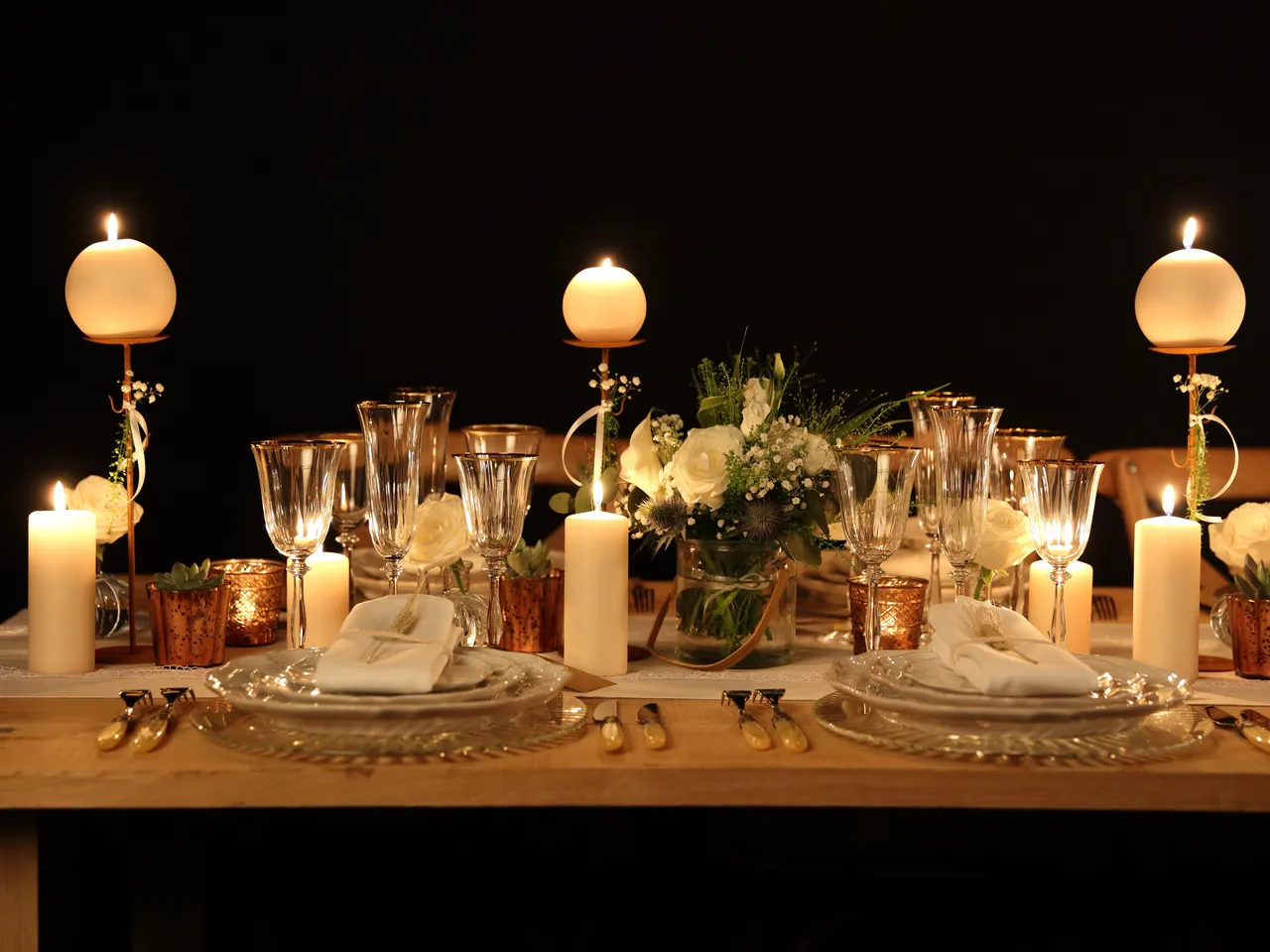 Elegant formal dinner table setting with white plates, crystal glasses, white napkins, candles in gold holders, white flowers in a central arrangement, and decorative gold accents on a wooden table against a dark background.