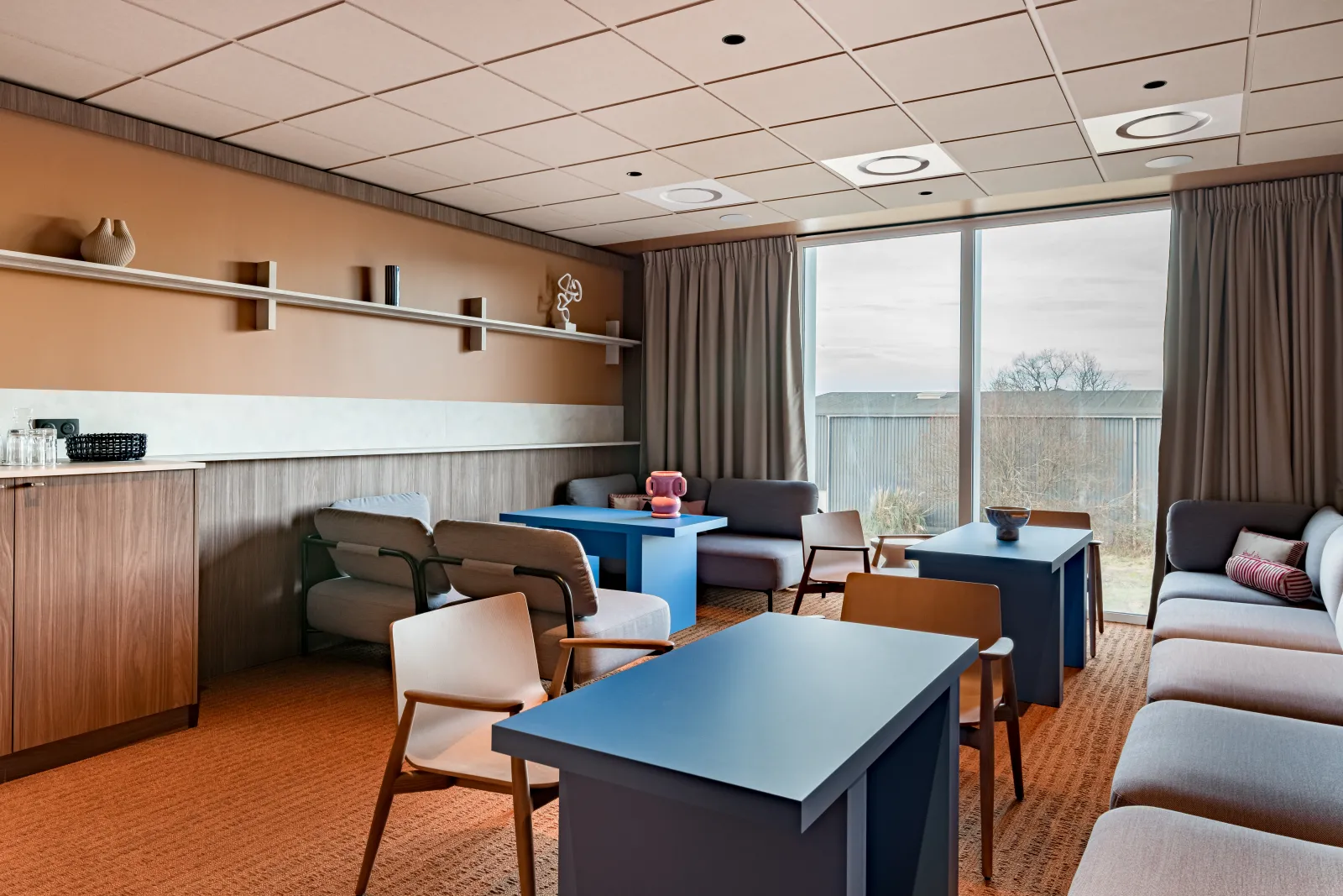 Modern lounge area at Hampton by Hilton Bordeaux Mérignac featuring blue tables, comfortable seating, orange carpet, and large windows overlooking the landscape