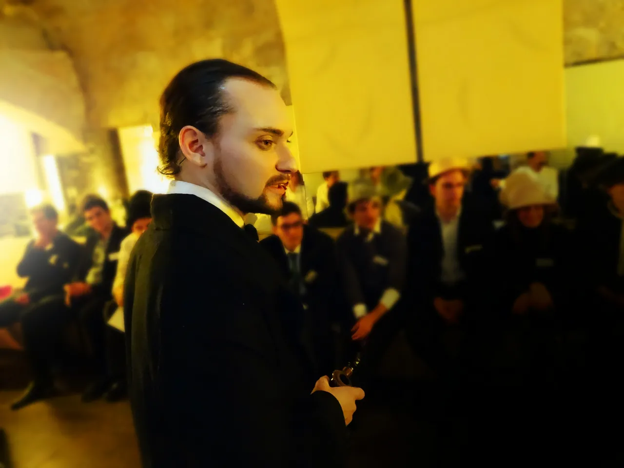 Man with dark hair and beard wearing a black jacket speaking to a crowd in a yellow-lit indoor venue in Bordeaux