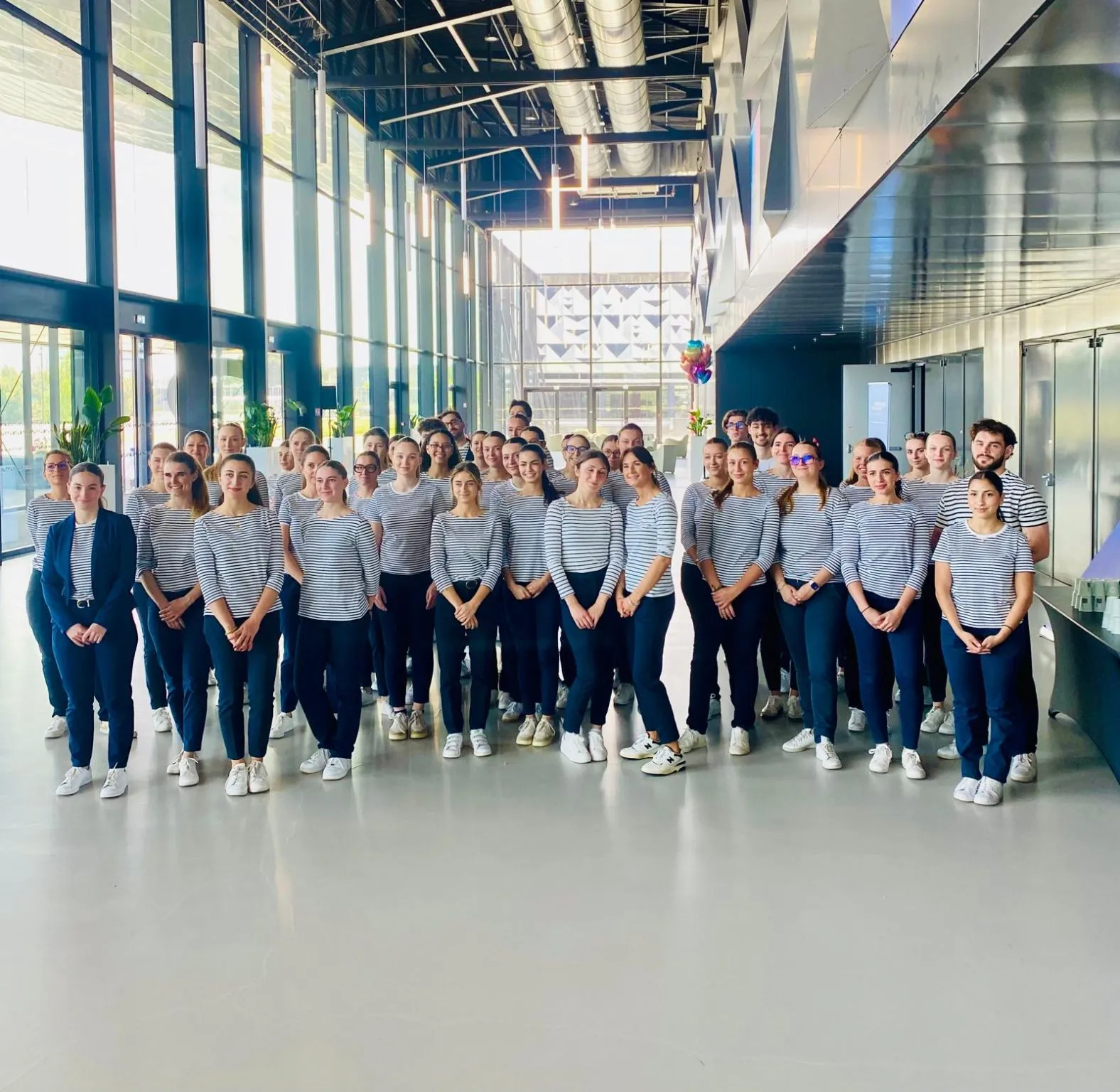 Large group of people wearing matching striped shirts and dark pants posing together in a modern, bright corridor with floor-to-ceiling windows and industrial ceiling elements.