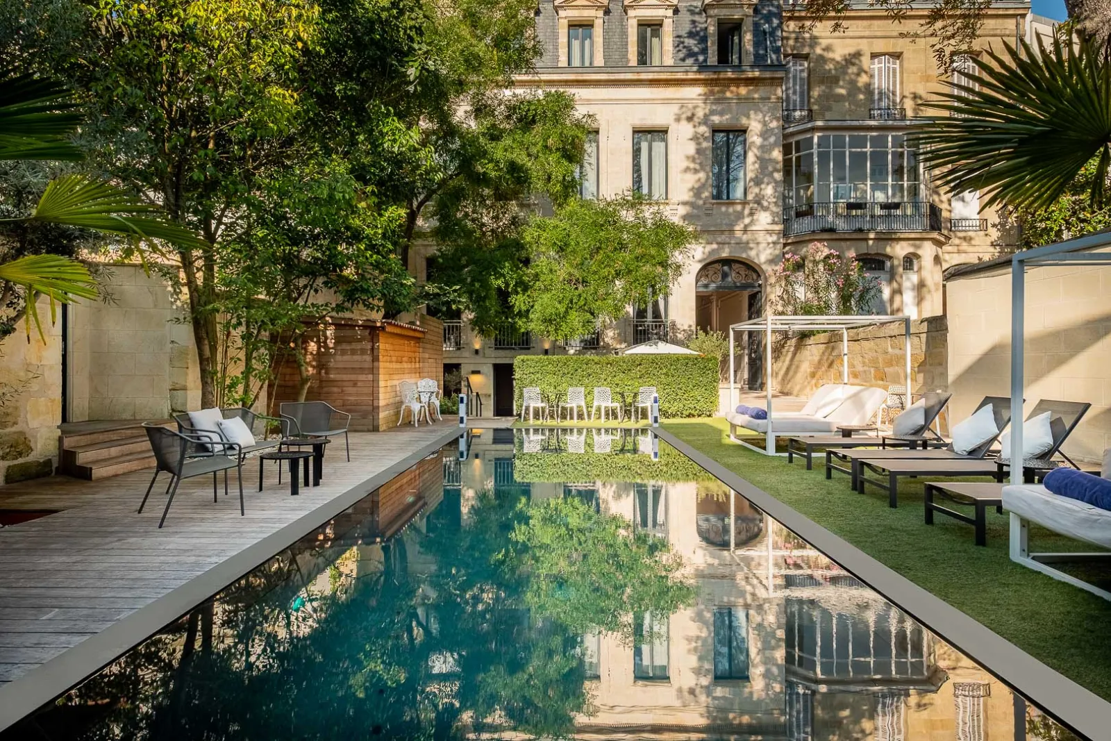 Luxury courtyard with a rectangular reflecting pool, lined with lounge chairs and sun beds, surrounded by manicured gardens and a historic Haussmann-style mansion in Bordeaux