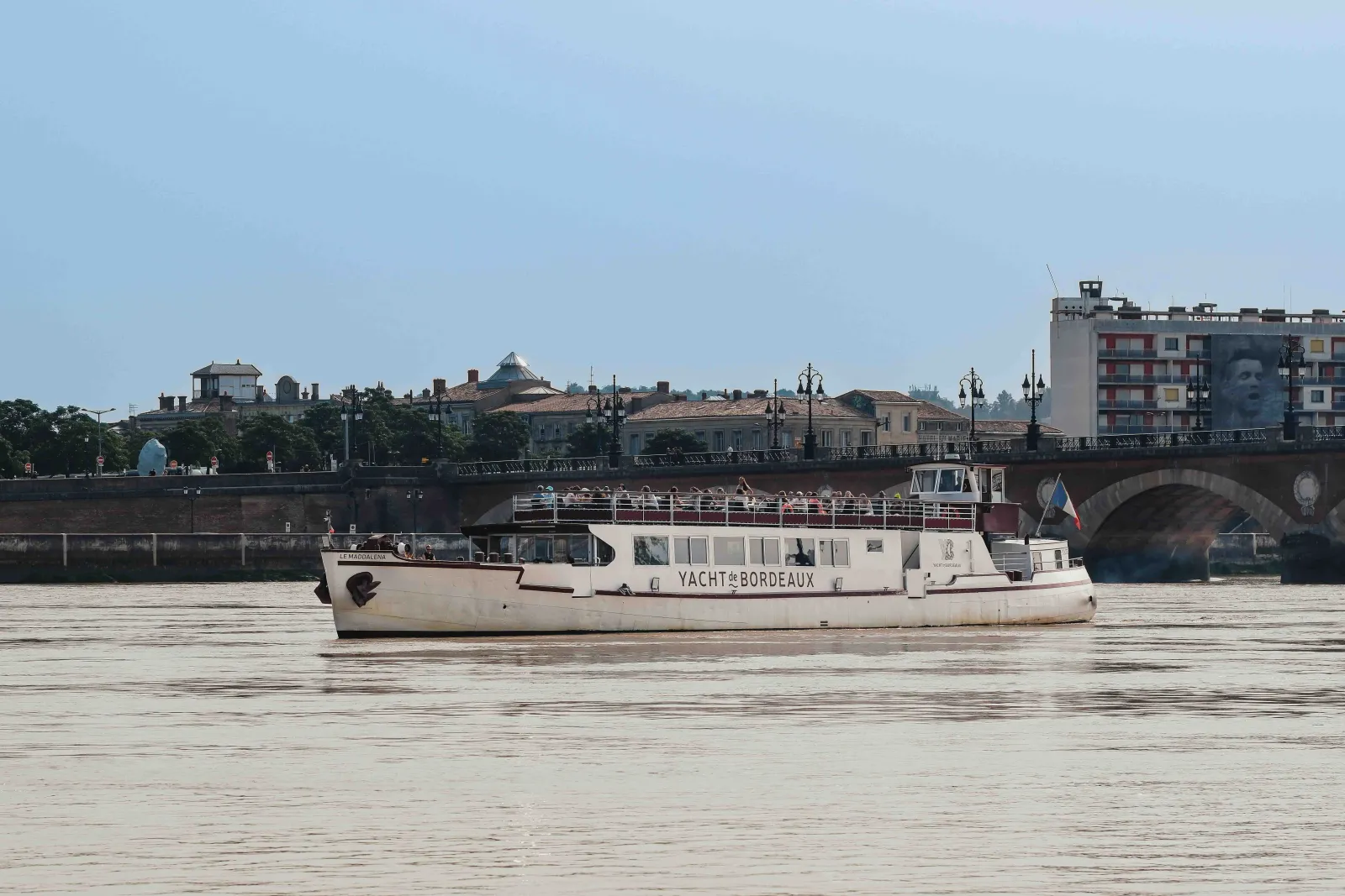 A white passenger river cruise boat named 'Yacht de Bordeaux' sailing on the Garonne River with historic Bordeaux buildings and a stone bridge in the background under a clear blue sky.