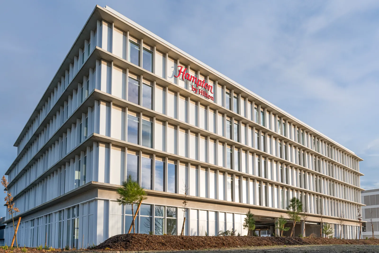 Modern Hampton by Hilton hotel building in Bordeaux Mérignac with contemporary architecture featuring white vertical louvers, multiple floors with large windows, and landscaped entrance.