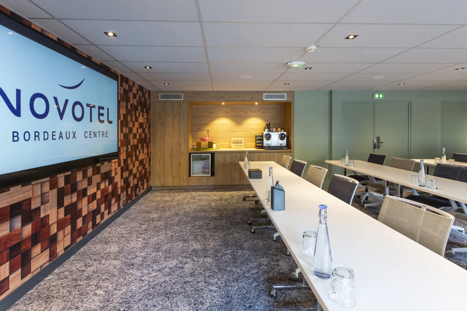 Modern meeting room at Novotel Bordeaux Centre hotel featuring a long white conference table with chairs, wooden wall paneling, projection screen displaying the hotel logo, and refreshment station with water and glasses.