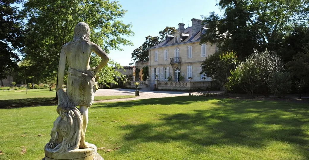 Stone statue of a female figure in the gardens of Château Kirwan in Bordeaux, with the 18th-century mansion visible in the background surrounded by trees