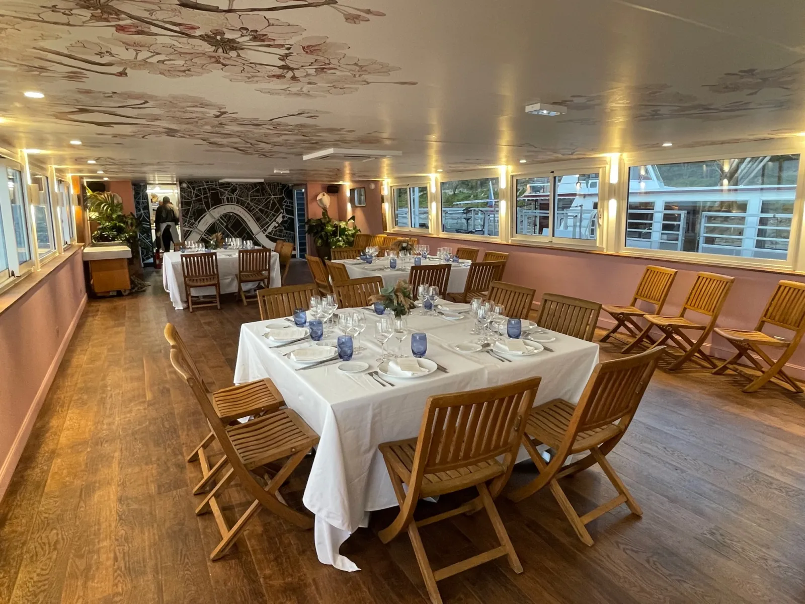 Elegant dining room on a luxury yacht with white tablecloths, wooden chairs, place settings, blue glassware, and floor-to-ceiling windows overlooking the city