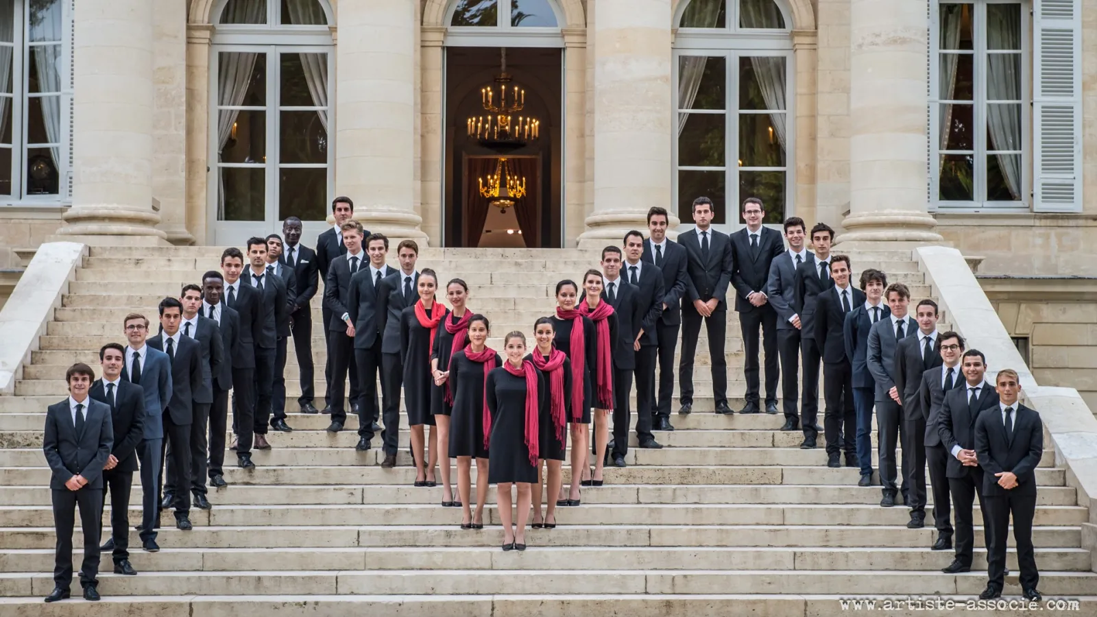 A formal group portrait of approximately 40 young professionals dressed in dark business suits and formal attire, with a central group of women wearing red scarves, positioned on the grand stone steps of an elegant neoclassical building with columns and o