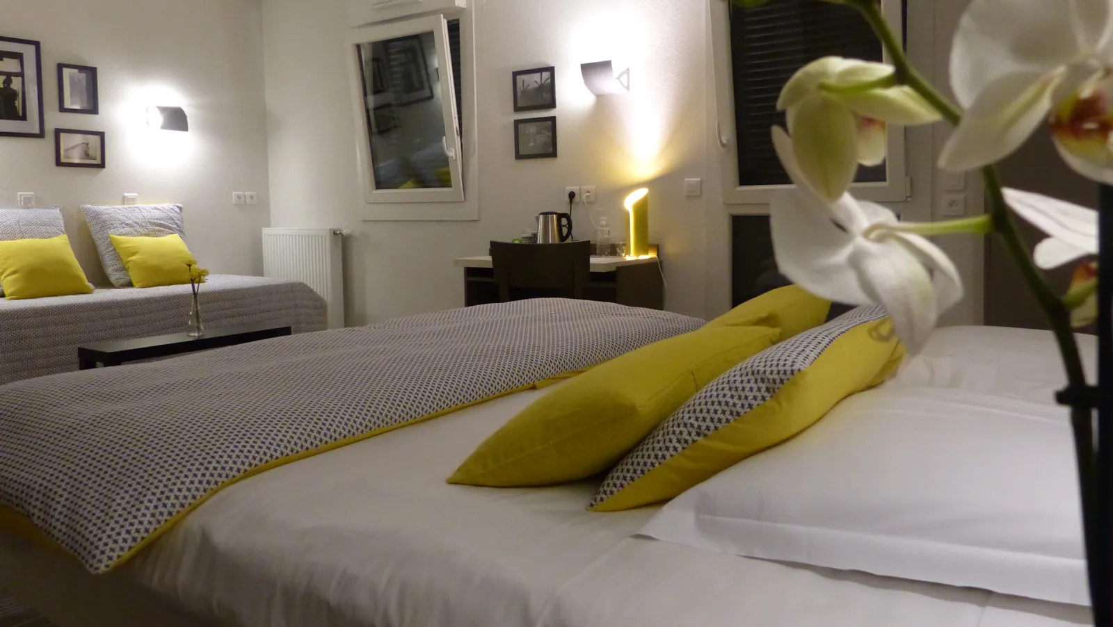 Modern bedroom at Aérocampus Aquitaine in Bordeaux featuring a bed with yellow and gray bedding, white pillows, fresh orchid flowers on the nightstand, a sofa with yellow cushions, framed artwork on walls, and contemporary lighting fixtures.