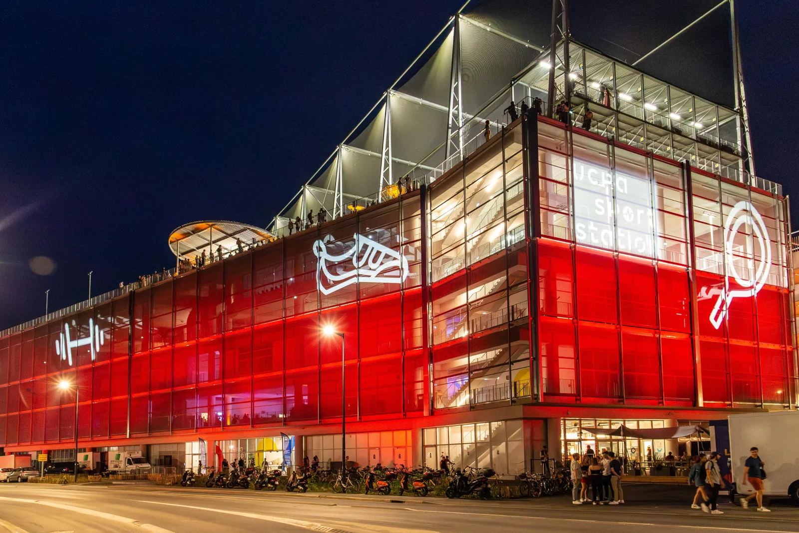 Night view of the UCPA Sport Station building in Bordeaux, featuring a modern red-lit facade with glass panels, metal framework, and illuminated upper levels with people visible on terraces.
