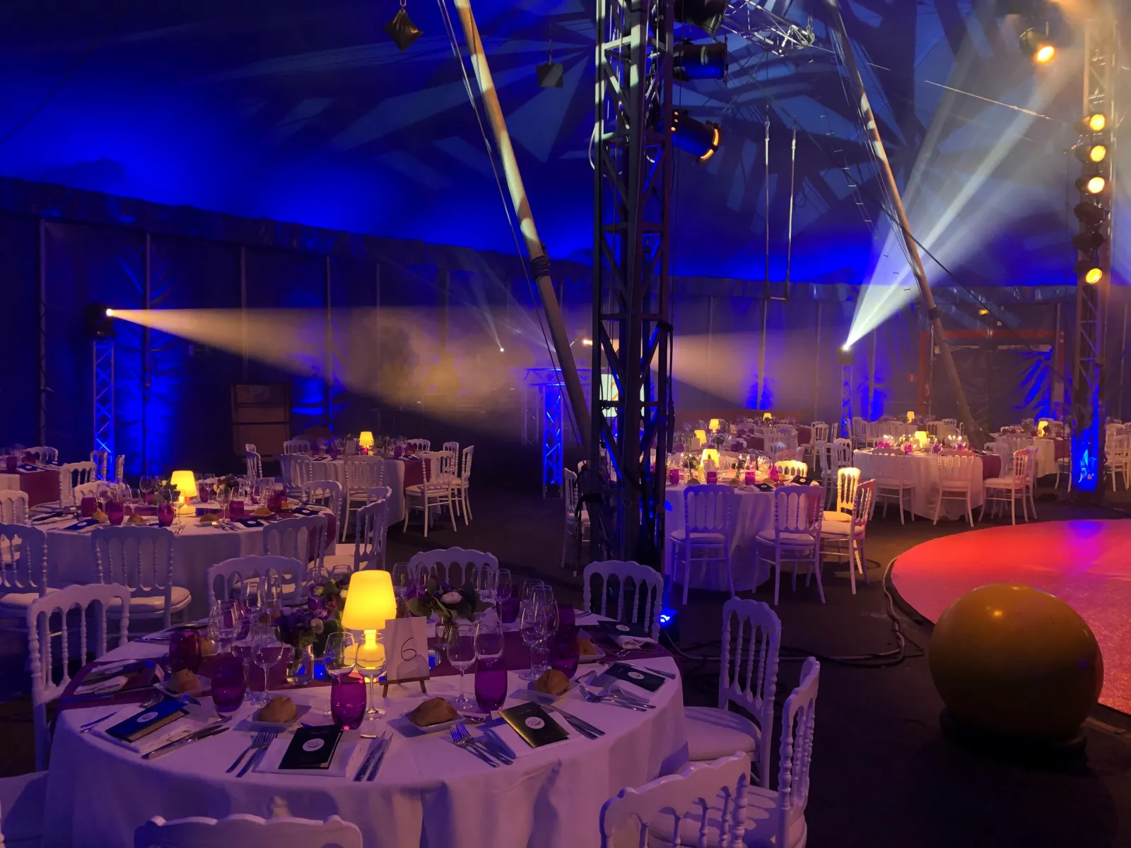 Elegant gala dinner setup in a large venue with round tables, white chairs, ambient blue and golden lighting, professional stage lighting rigs overhead, and a dance floor illuminated in red, ready for an evening event.