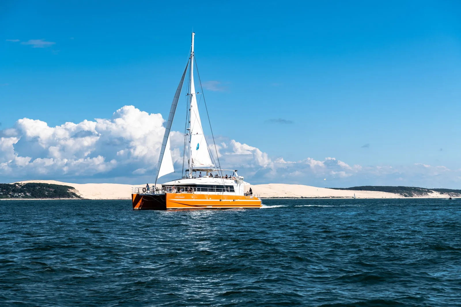 Orange and white catamaran sailboat with white sails deployed, sailing in blue waters near white sand dunes and coastal cliffs under a clear sky with white clouds