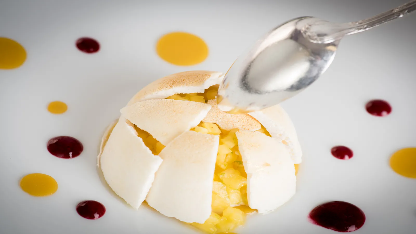 A broken meringue shell filled with custard cream, being drizzled with sauce, surrounded by yellow and dark red sauce dots on a white plate.