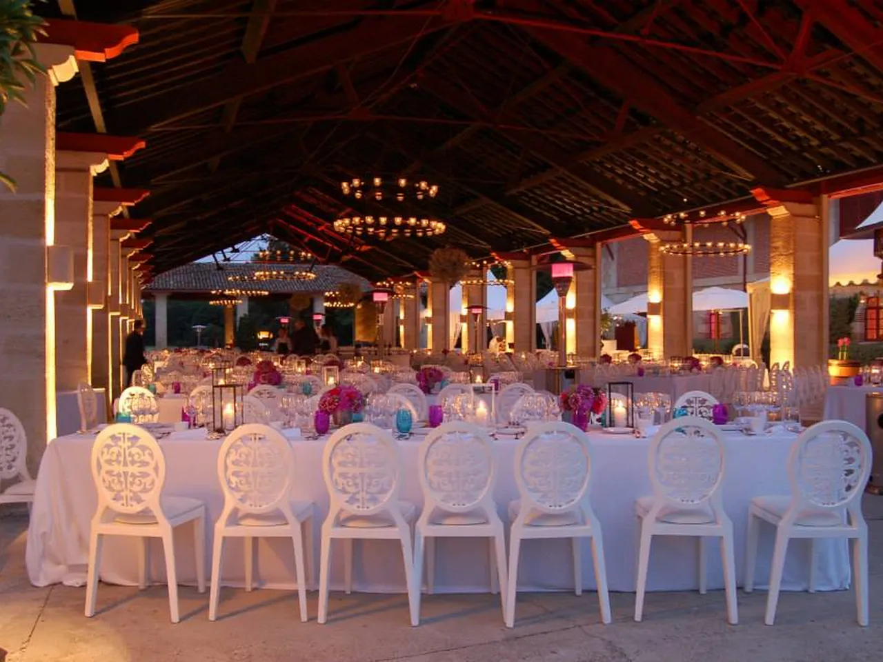 Elegant outdoor wedding reception venue at Château Giscours in Bordeaux, featuring white decorative chairs along long tables with floral centerpieces in pink and purple tones, under a covered pavilion with warm golden lighting at dusk.