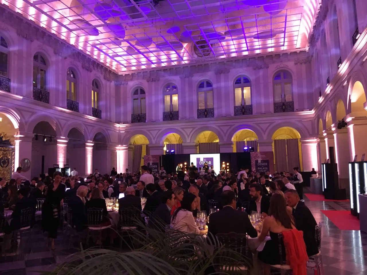 Elegant gala event in a grand Haussmann-style ballroom in Bordeaux with purple and pink lighting, featuring guests in formal attire seated at tables, ornate architecture with arched colonnades, and a stage with branding in the background.