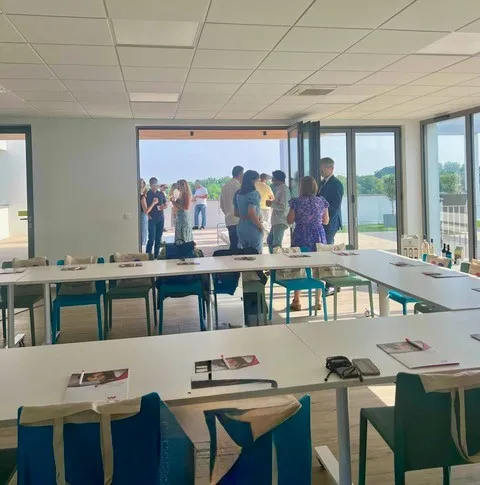 Modern interior space with floor-to-ceiling windows overlooking a waterfront landscape, featuring wooden tables with blue chairs and a group of people gathered on an adjacent terrace.