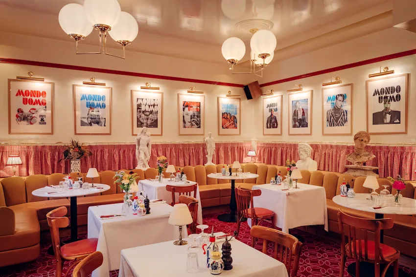 Elegant dining room at Osteria Palatino in Bordeaux featuring red and white checkered banquette seating, round tables with white tablecloths, vintage magazine covers on walls, and warm amber lighting fixtures.