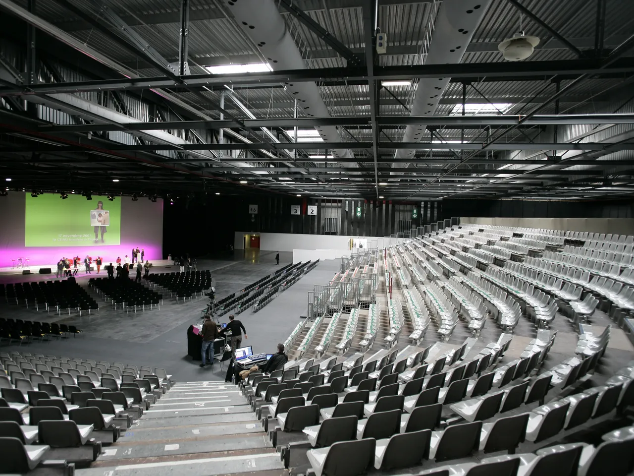Large modern exhibition hall with rows of white seats facing a stage with pink lighting where a presentation is taking place, featuring industrial ceiling with metal beams and skylights.