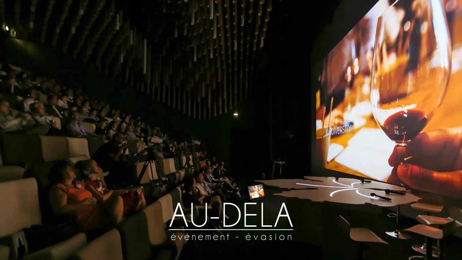 Indoor event venue with audience seated in tiered rows watching a large projection screen displaying amber-colored lighting and biodiversity content, with Au-Dela event company branding visible