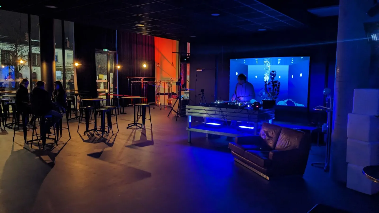 Modern event space at L'Estrade in Bordeaux with blue ambient lighting, DJ booth with screen display, leather seating, bar tables, and visitors in a contemporary venue setting.