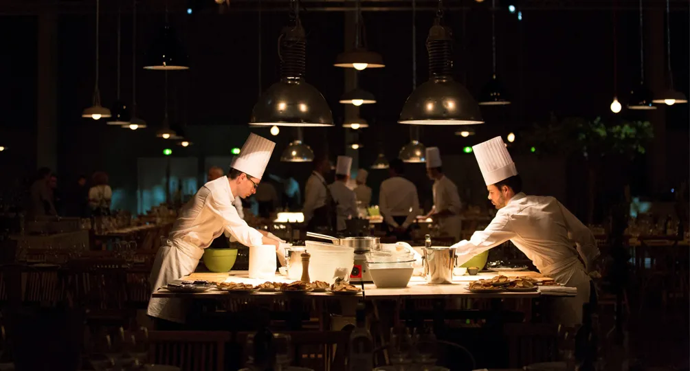 Two chefs in white uniforms and chef hats prepare food at a counter in a modern restaurant kitchen with hanging industrial lights, while diners are visible in the background.