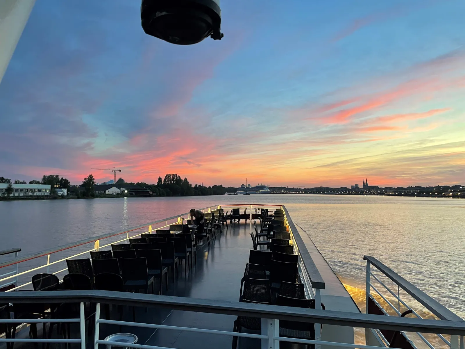 Upper deck of a modern yacht with empty black chairs arranged in rows, sailing on a calm river at sunset with a city skyline and church spires visible in the background