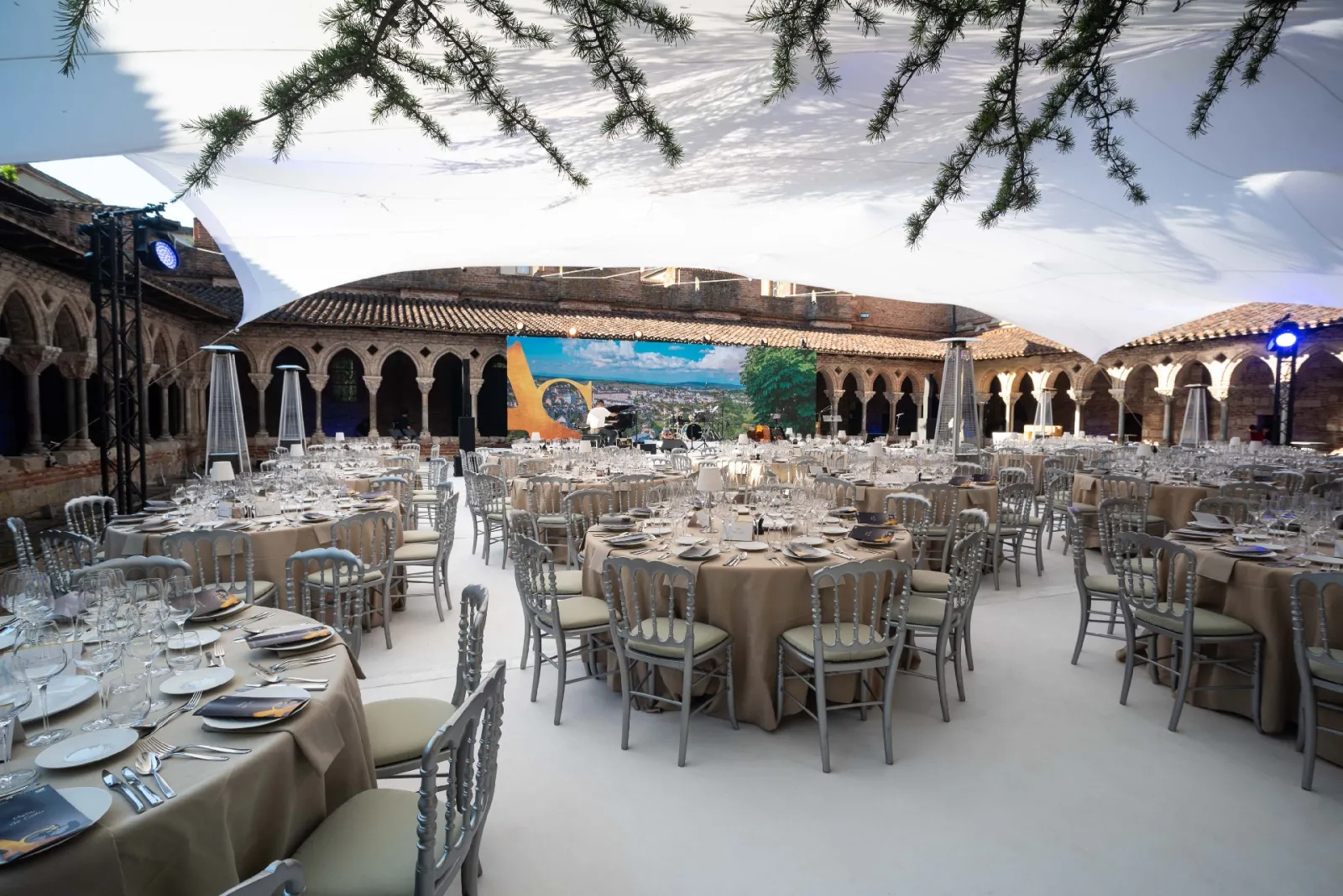 Elegant outdoor banquet setup in a historic courtyard with stone arches, round tables with beige linens, chiavari chairs, and formal place settings under a blue sky