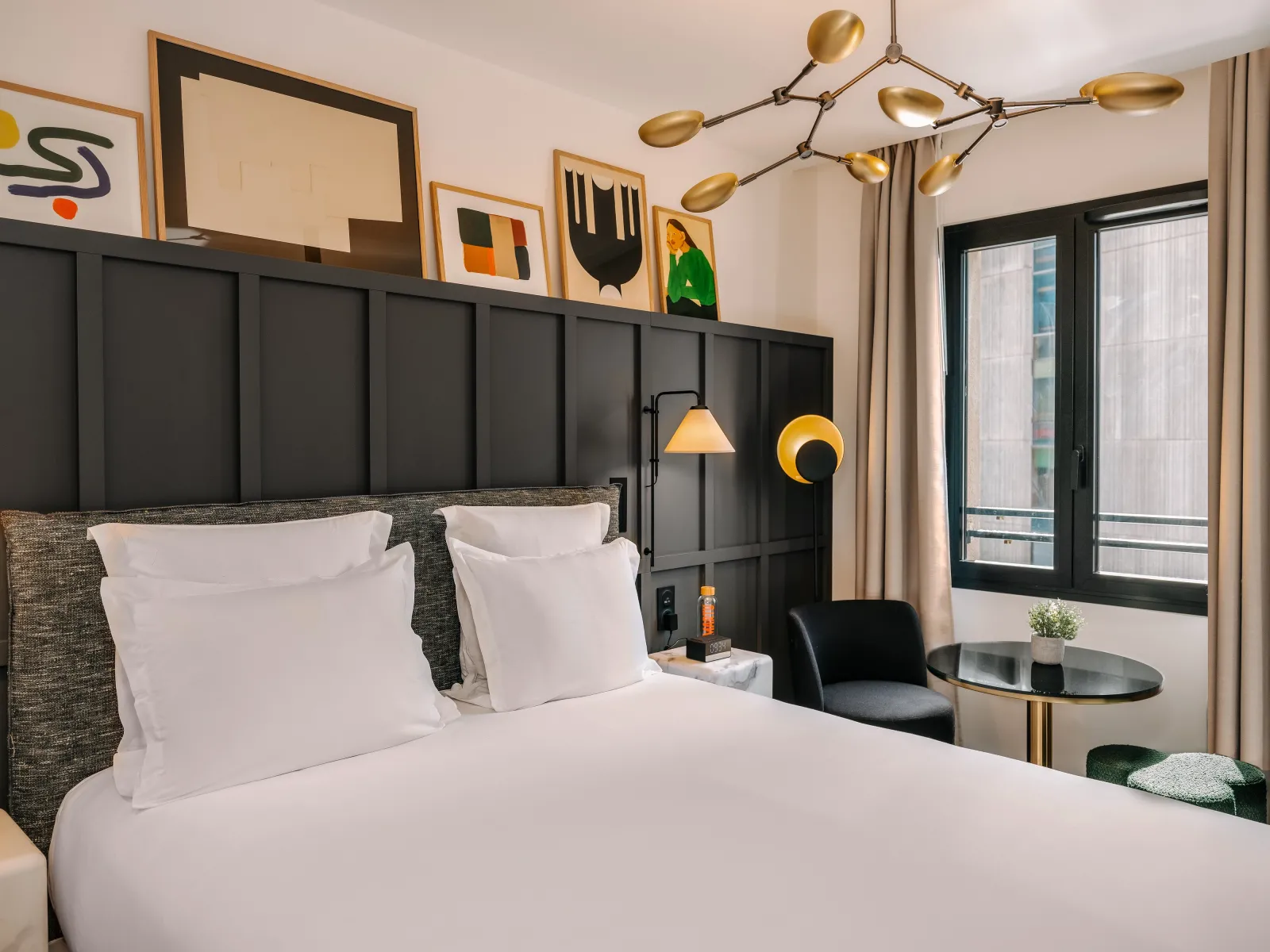 Modern hotel bedroom at Marty Hôtel Bordeaux featuring a white bed with gray upholstered headboard, brass pendant lighting, black accent wall with framed artwork, and glass doors opening to a balcony