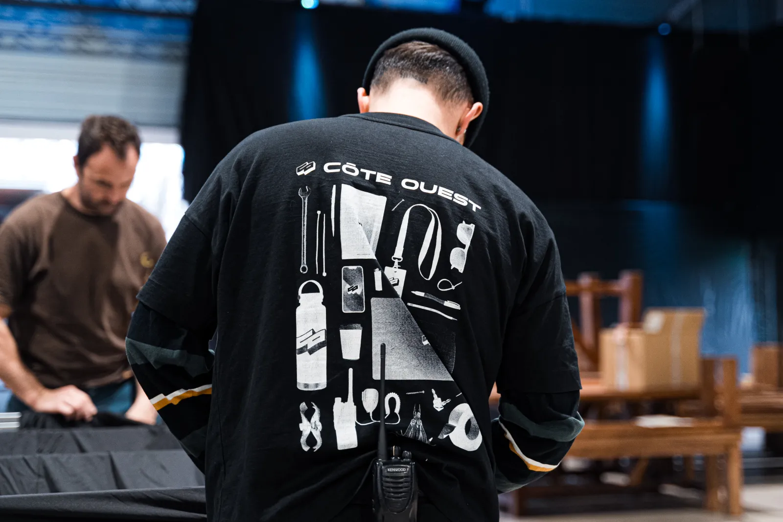 Person wearing a black 'Côte Ouest' branded jacket displaying outdoor and camping gear icons on the back, standing in an indoor workshop or event space with another person visible in the background.