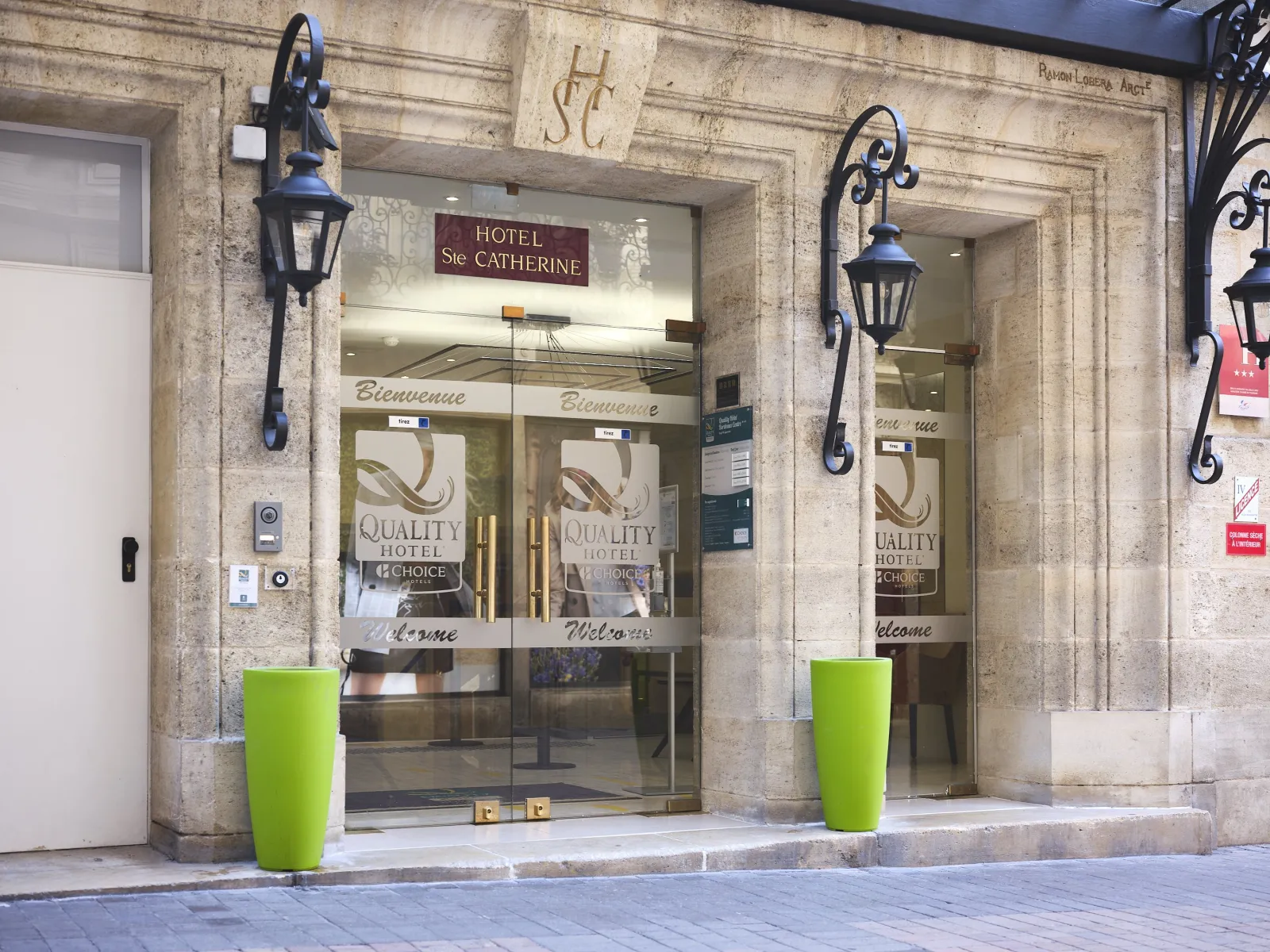 Entrance of Hotel Sainte Catherine, a Quality Hotel in Bordeaux city center, featuring glass doors, decorative wrought iron wall lamps, and bright green cylindrical planters on the stone facade.