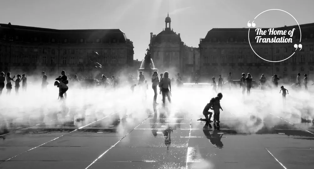 Black and white photograph of a public square in Bordeaux with fountains spraying water, people playing and walking through the mist, historic buildings and a domed structure in the background.