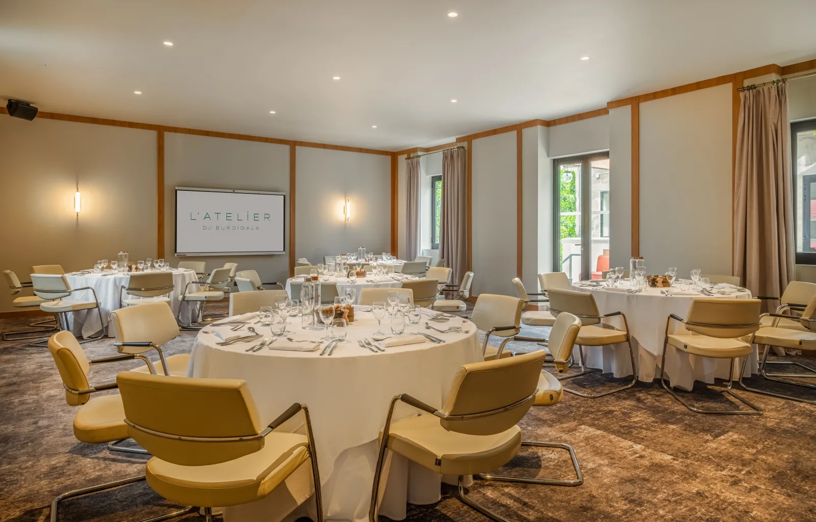 Elegant banquet hall at Hotel Burdigala featuring round tables with white tablecloths, yellow modern chairs, wooden beam details, soft lighting, and a presentation screen displaying 'L'Atelier du Burdigala'.