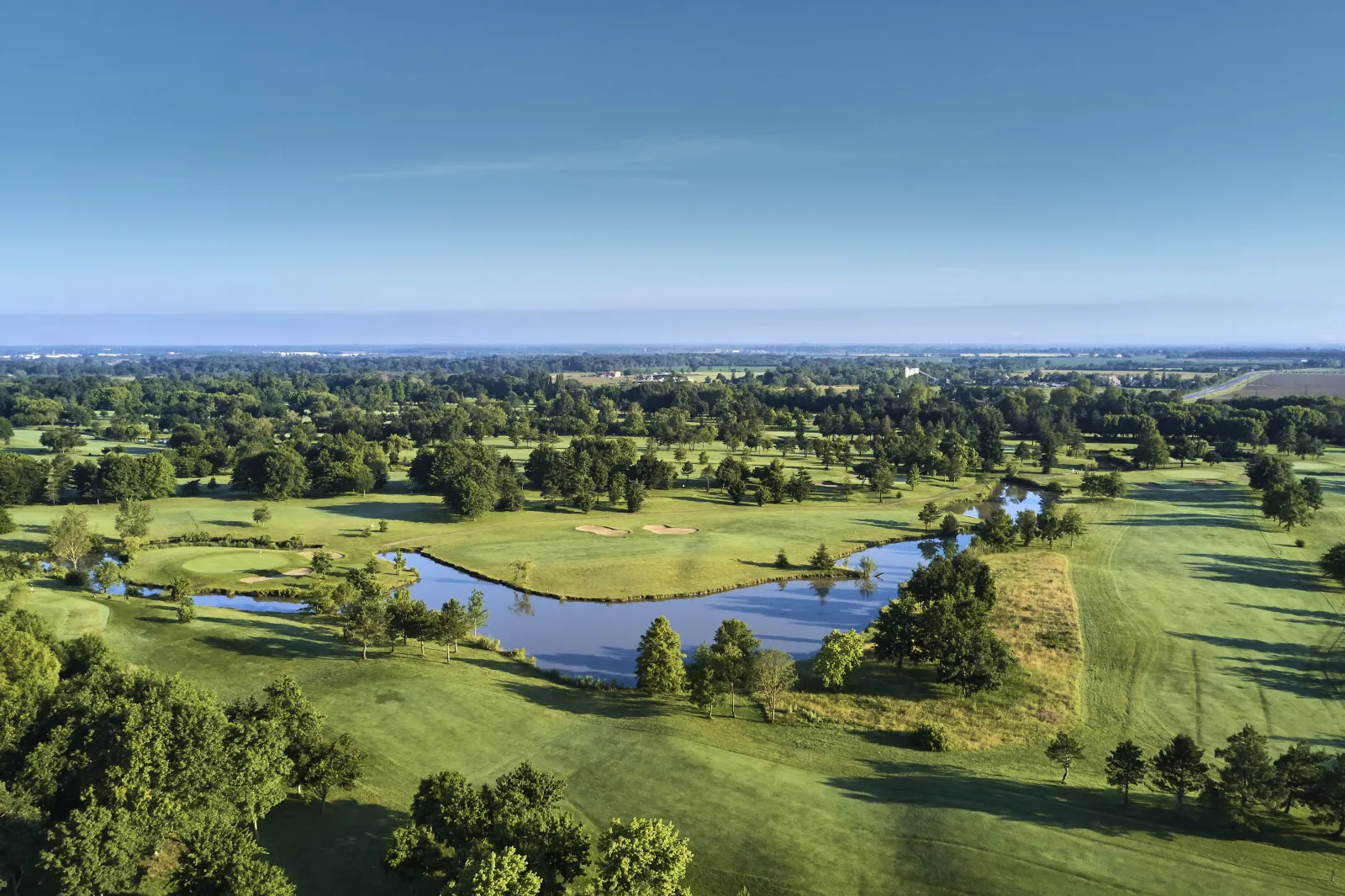 Aerial view of Bordeaux Lac Golf course featuring manicured greens, scattered trees, and a winding blue water hazard under clear skies