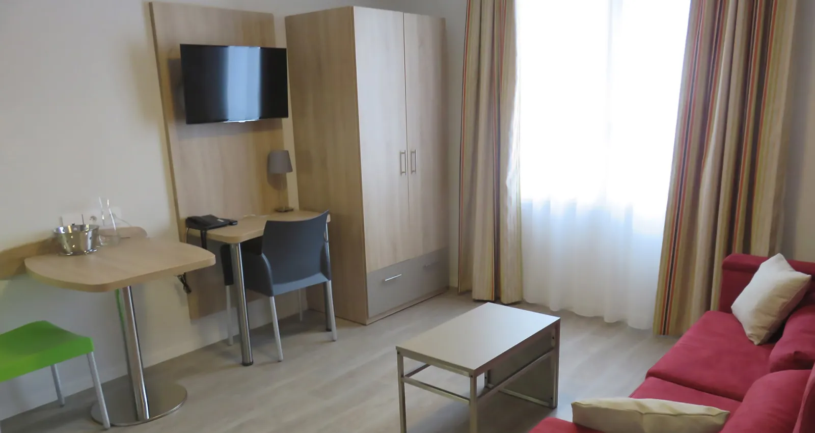 Modern hotel room with red sofa, work desk with TV, wardrobe, coffee table, and window with curtains overlooking the city