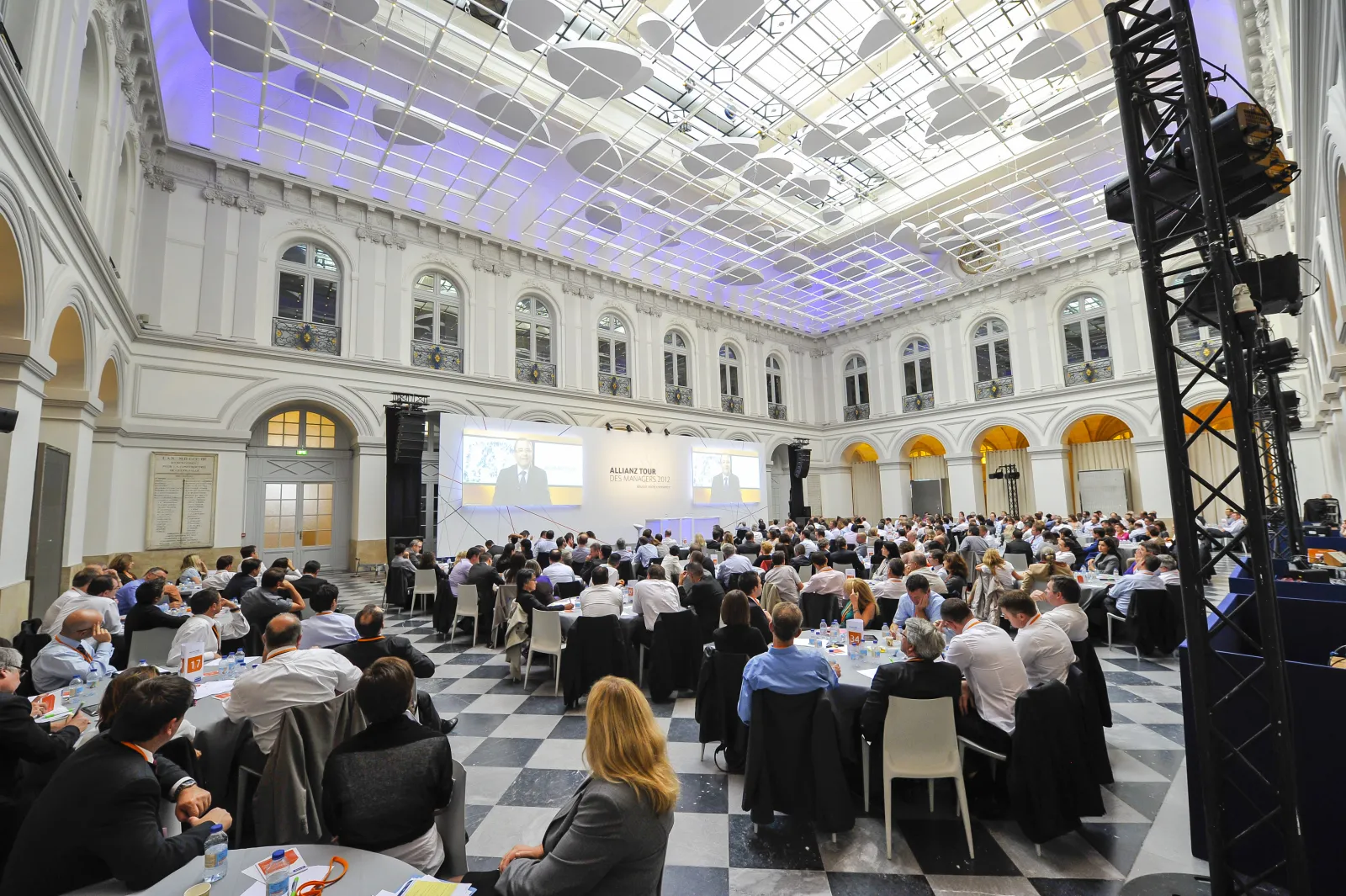 Large formal conference or banquet event in the neoclassical Palais de la Bourse in Bordeaux, with hundreds of attendees seated at round tables, a stage with screens displaying a presentation, decorative purple and white lighting on the glass ceiling, and
