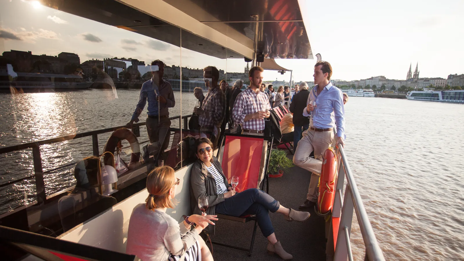 Passengers enjoying a river cruise on a modern boat with a glass-enclosed upper deck, sailing on the Garonne River in Bordeaux with the city skyline and cathedral visible in the background.