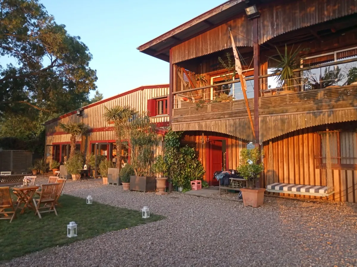Modern industrial-style building with corrugated metal exterior in warm orange and red tones, featuring wooden balconies and terraces with potted plants, gravel courtyard, and green lawn area at dusk.