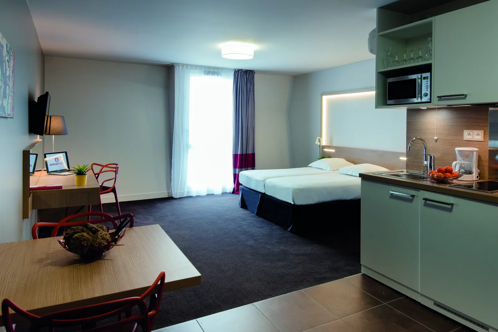 Modern studio apartment at Appart City Confort Bordeaux Chartrons featuring a kitchenette with green cabinets, dining area with red chairs, work desk with laptop, queen bed with white linens, and large window with purple curtains.