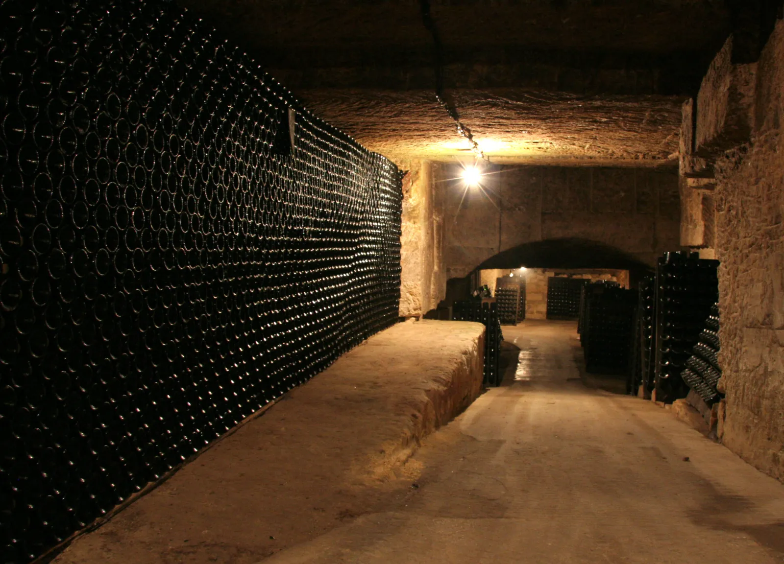 Underground wine cellar in Bordeaux with rows of black wine bottles stored in racks along the stone walls, illuminated by a single overhead light