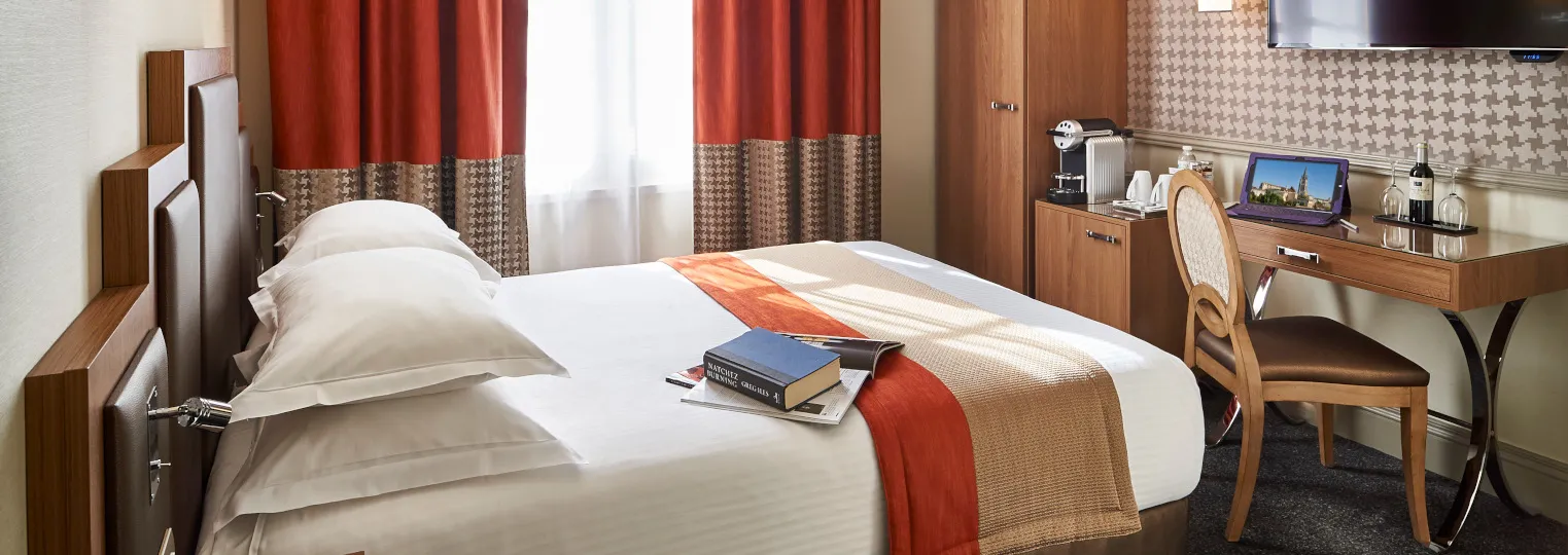 Modern hotel bedroom with a queen bed, white linens, red and beige decorative accents, wooden furniture, desk with chair, curtained window, and flat-screen TV mounted on the wall