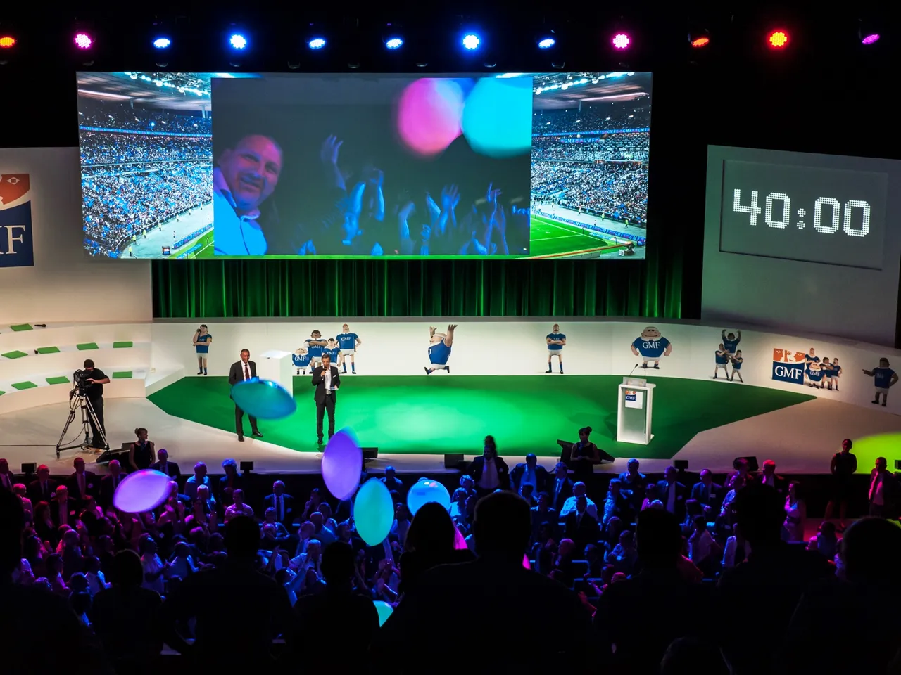 Large conference stage with green football field design, illuminated with colorful lighting, displaying large screens showing stadium crowd and sports content, with audience in foreground and speakers on stage at Bordeaux Congress Centre