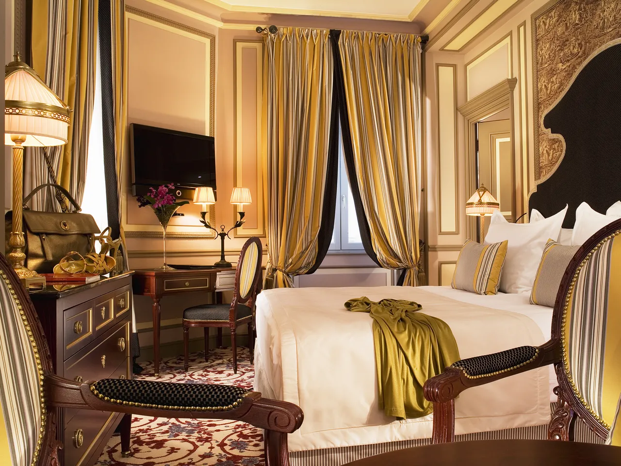 Luxurious hotel bedroom at InterContinental Grand Hôtel de Bordeaux featuring a bed with striped upholstery, gold and cream drapes, dark wood furniture, ornate mirrors, and elegant classical décor.