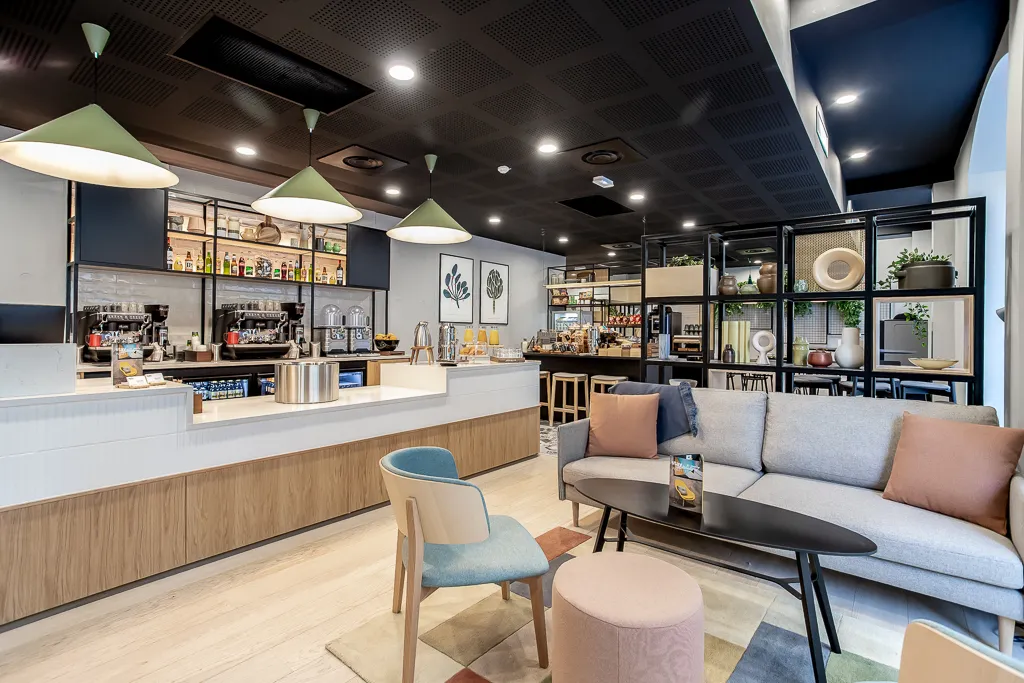 Modern aparthotel lounge and café area with white counter, wooden accents, comfortable seating, shelving displays, and contemporary lighting at Staycity Bordeaux City Centre