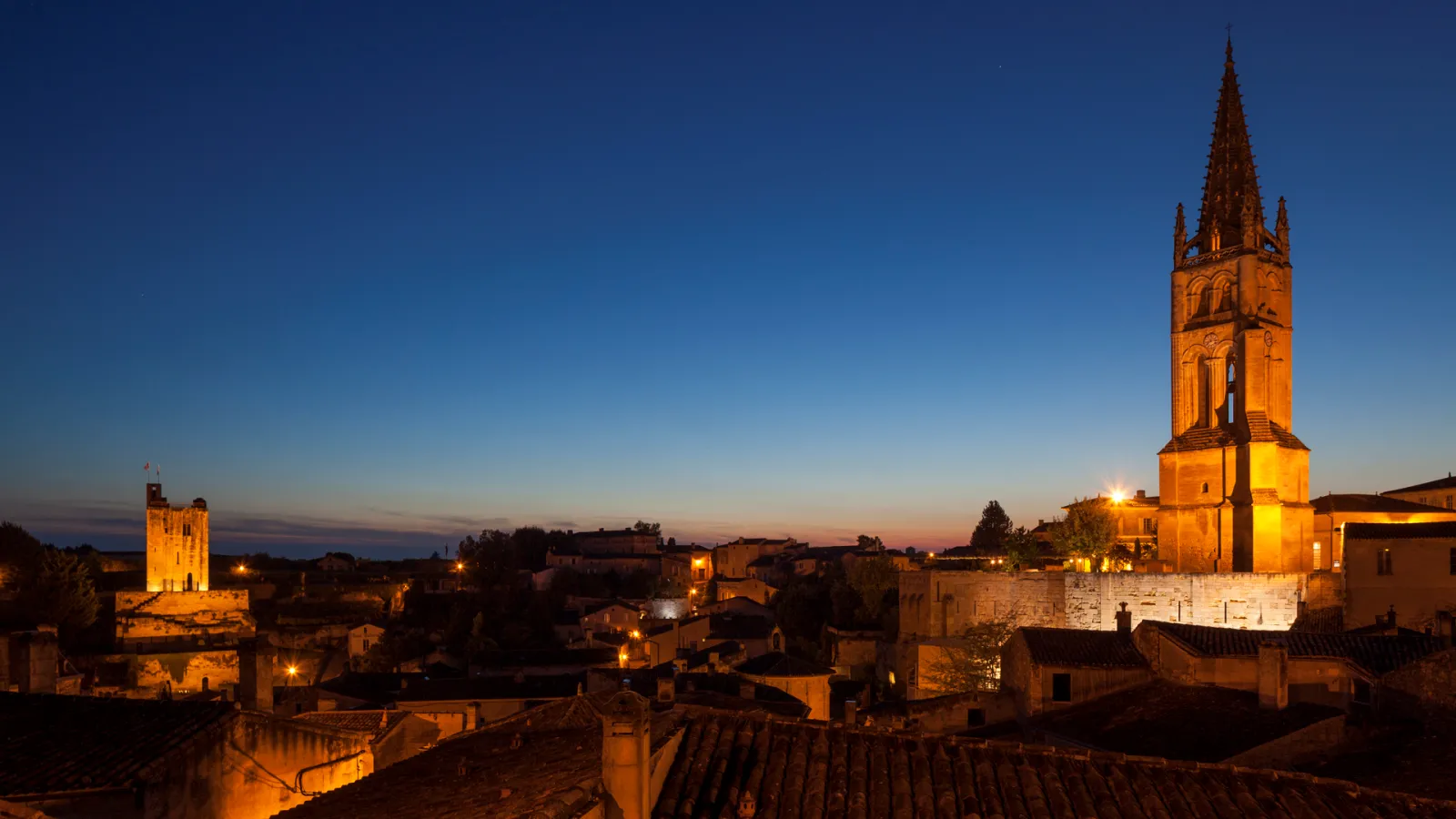 Night view of a historic medieval town with illuminated stone buildings and a tall church tower with spire, located in the Saint-Emilionnais region near Bordeaux.