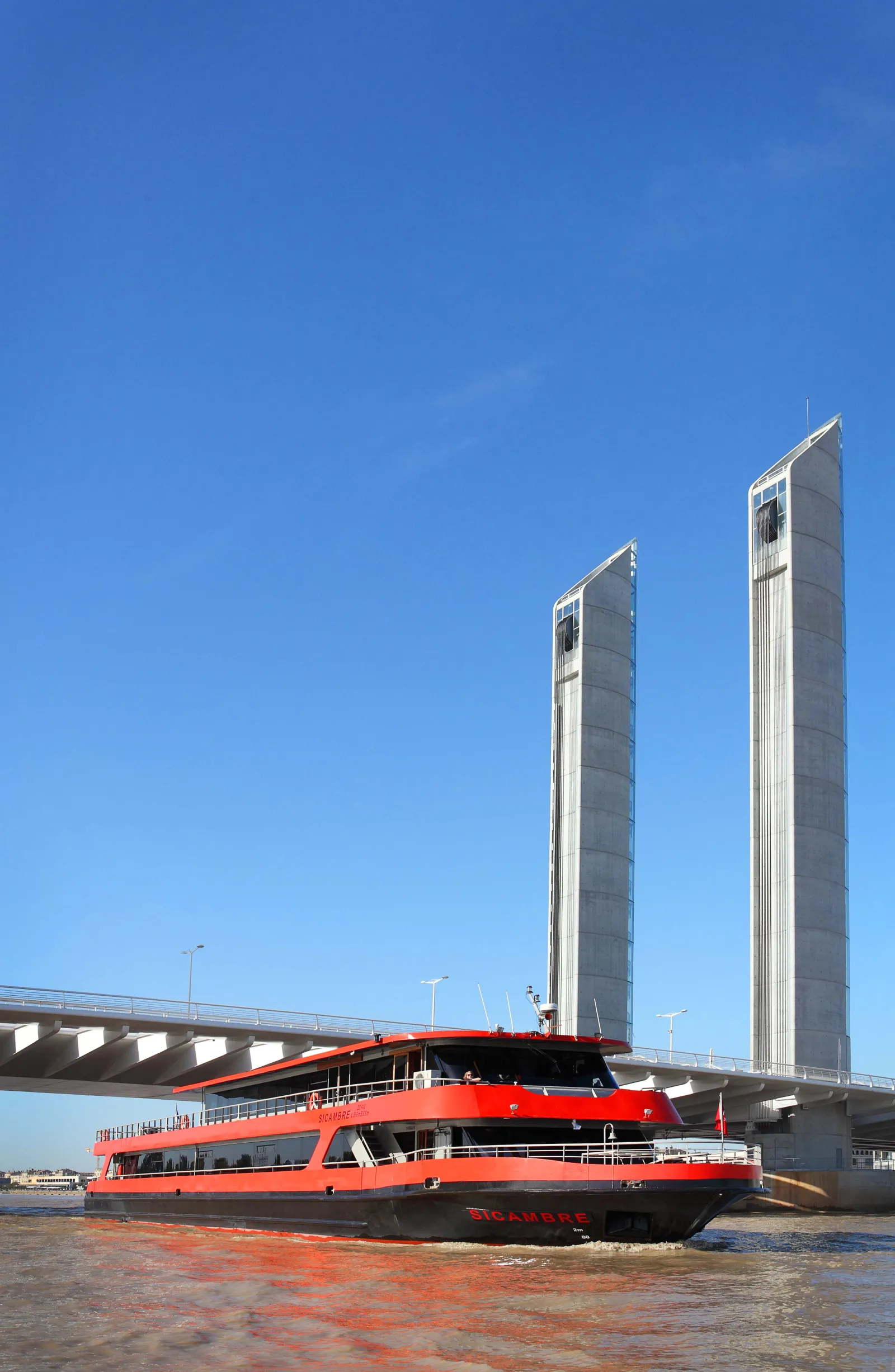 Red and black passenger ferry boat moored on the Garonne River in Bordeaux, with two modern twin towers and a bridge in the background under a clear blue sky.