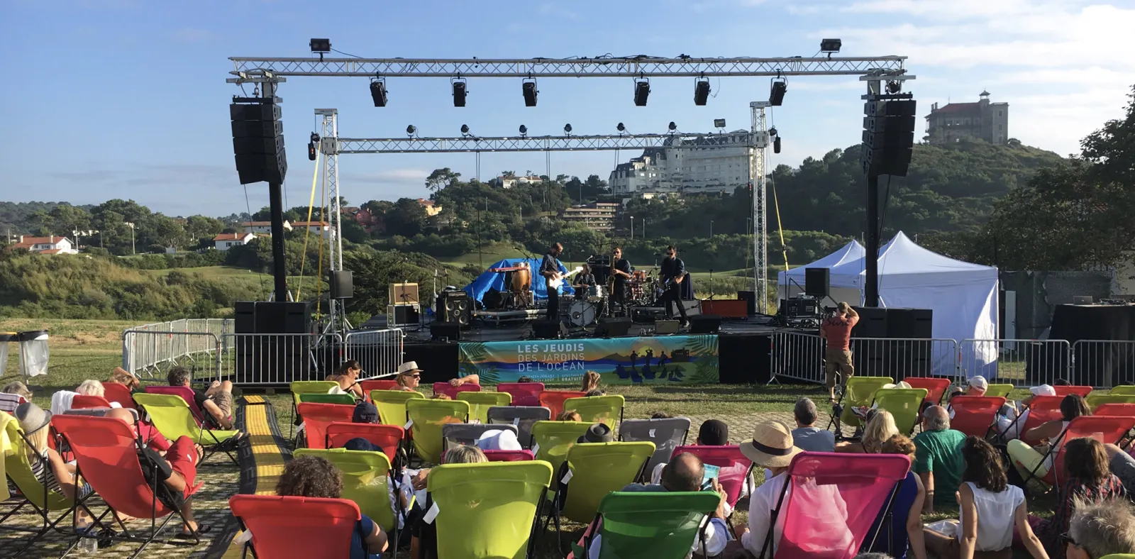 Outdoor concert event in Bordeaux with a band performing on stage under professional lighting rigs, surrounded by colorful seating and audience members, with hillside cityscape in the background